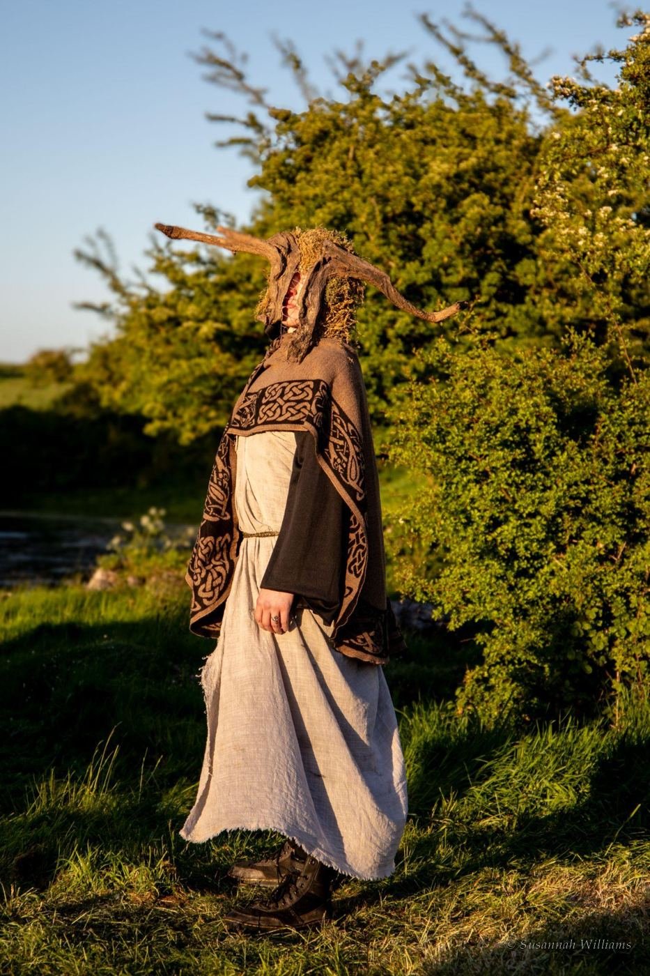 A figure in a handmade antlered headdress and woven cloak, backlit at dusk.