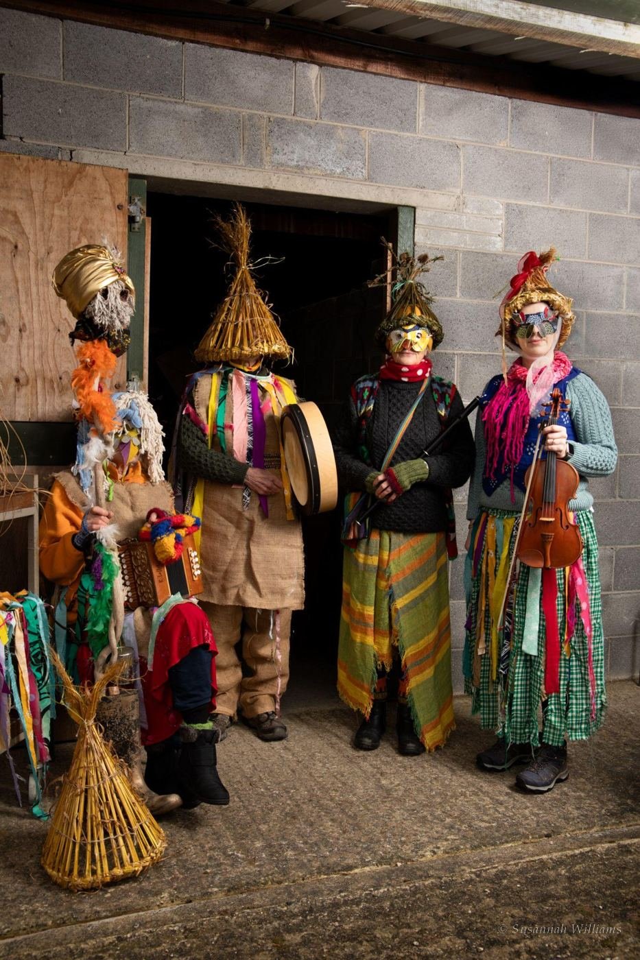 Four Mummers in straw and rag costumes in a barn doorway with a drum, concertina and fiddle.