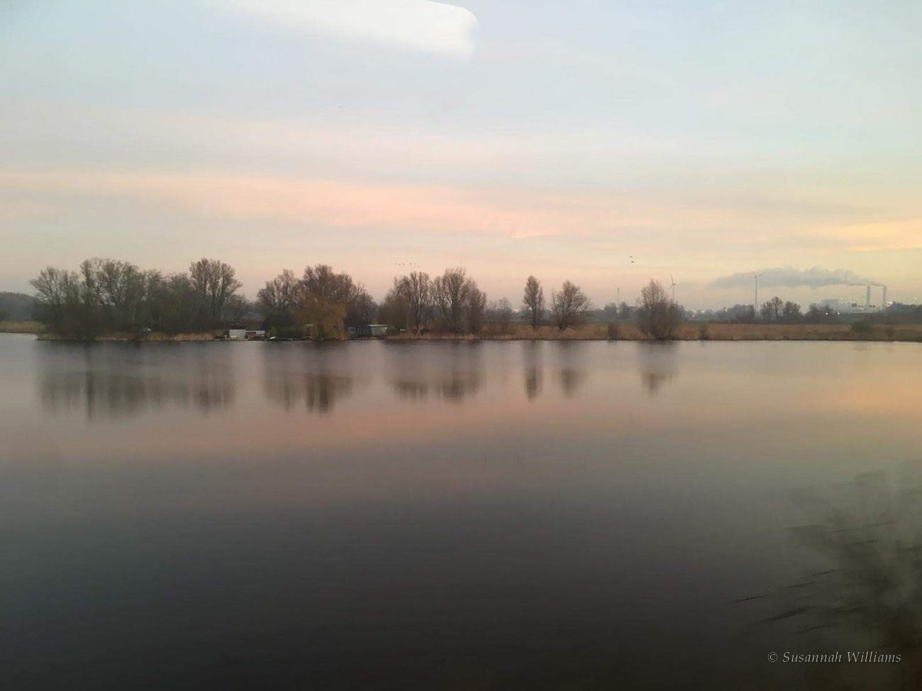 A still lake at dusk with bare trees reflected in the water.