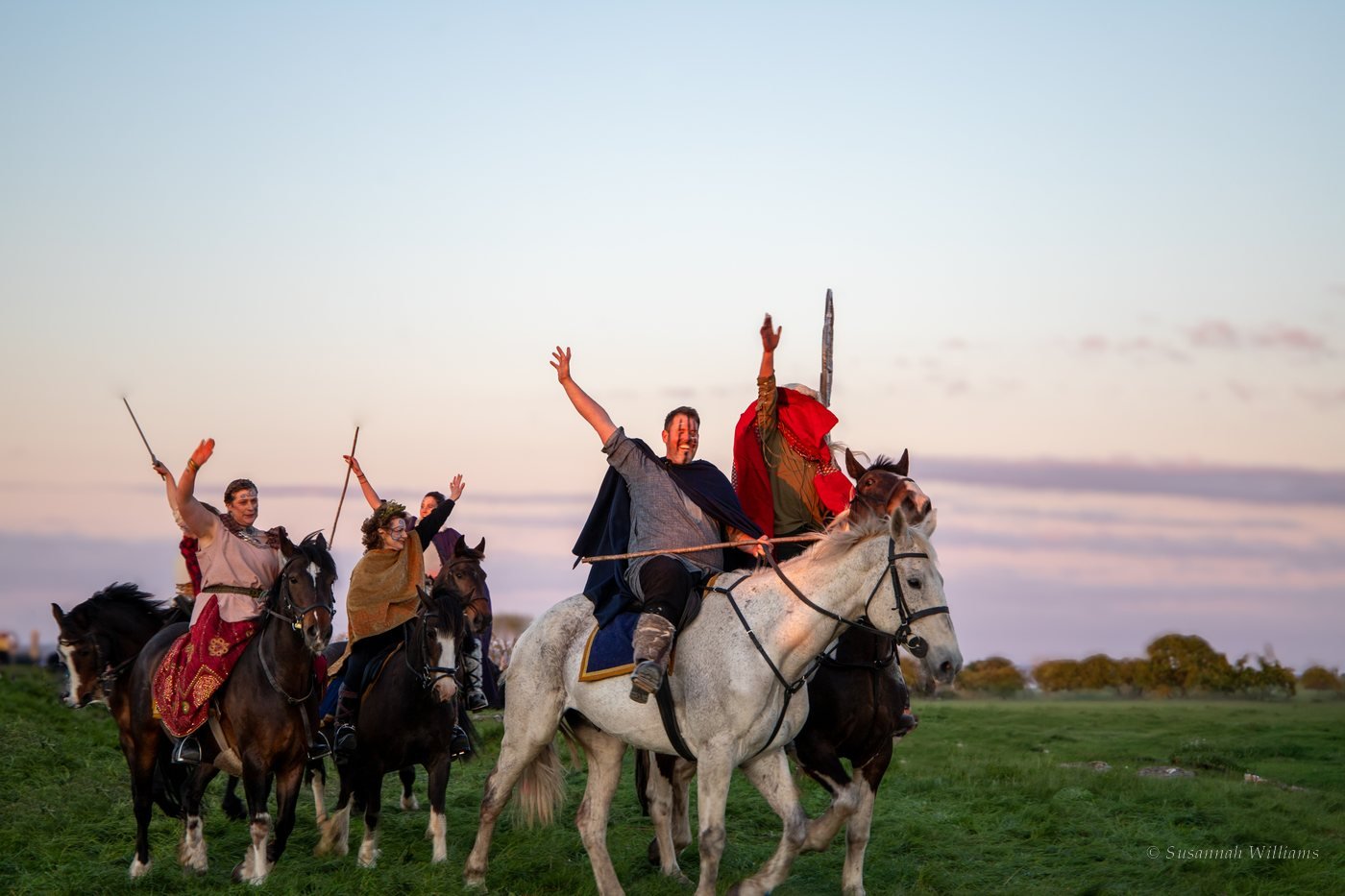 Riders in Celtic costume on horseback, arms raised in celebration against a pink evening sky.
