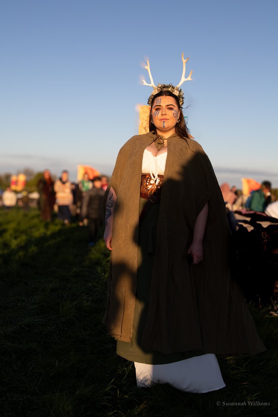 A woman crowned with white antlers and flowers, blue markings painted on her face, standing in golden evening light.