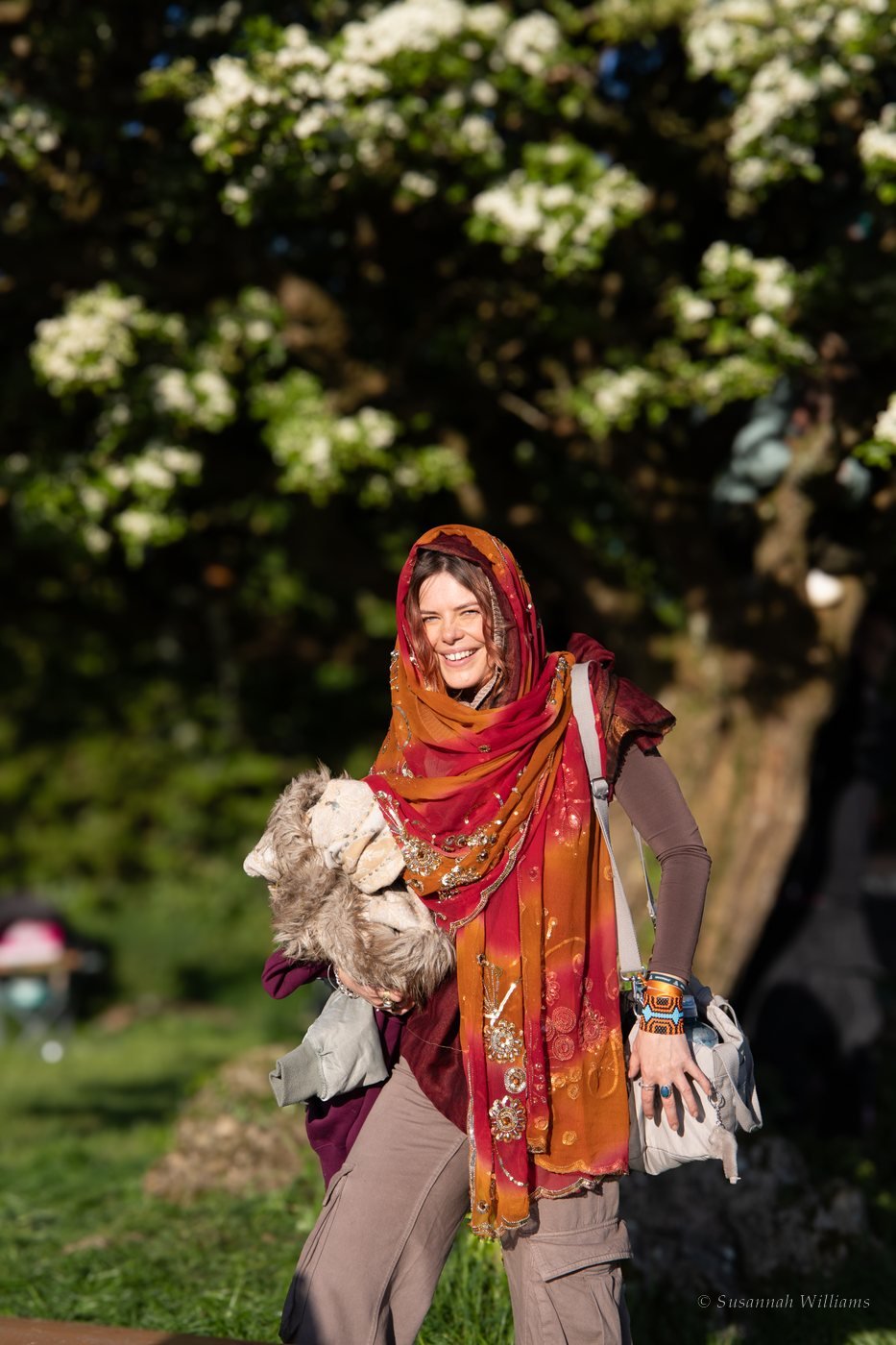 A smiling woman in a red and orange sequinned scarf walking beneath a hawthorn tree in full white bloom.