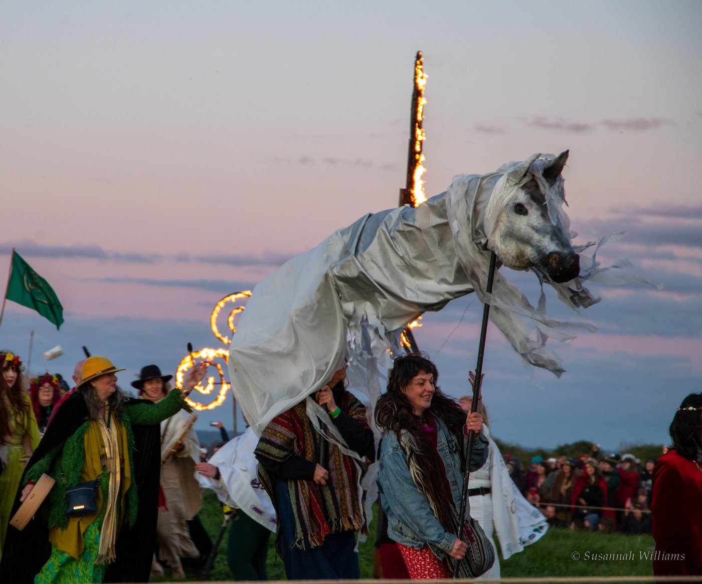 A large white horse puppet carried through the crowd, fire spirals burning behind it at dusk.