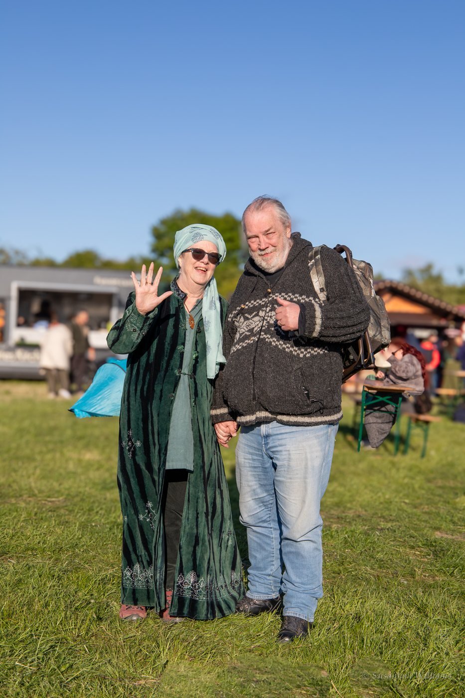 An older couple holding hands, she waving in a long green velvet coat, he giving a thumbs up.