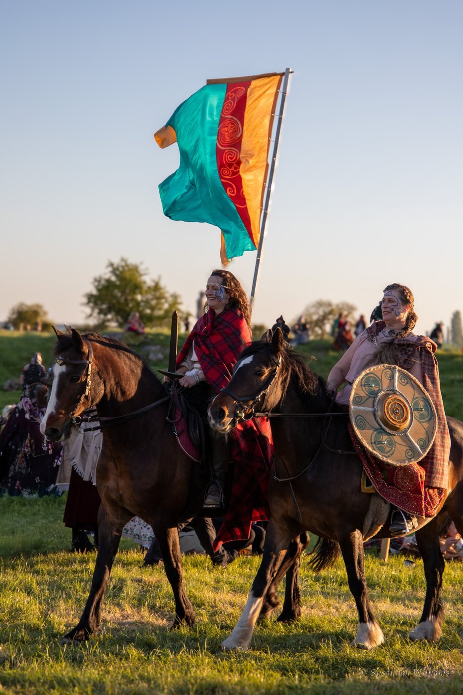 Two women on horseback with painted faces, a shield and a sword, a silk banner flying above them.