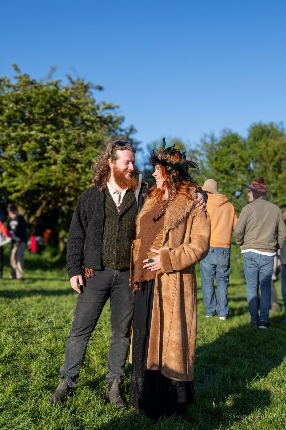 A couple smiling at each other, she in a feathered headdress and long sheepskin coat.