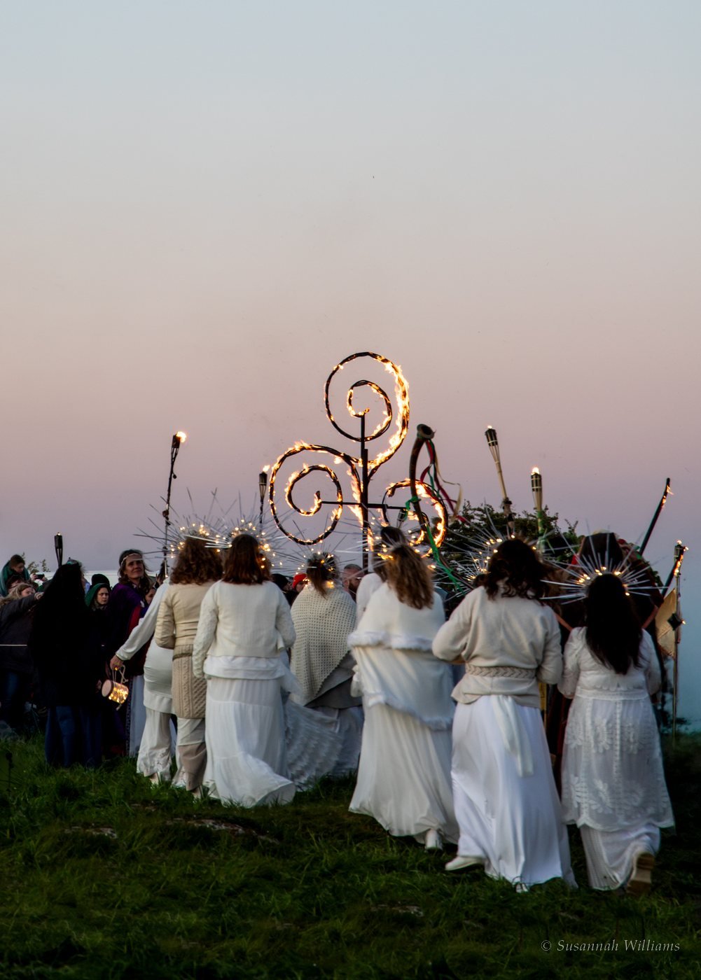 Women in white with crowns of light walking towards burning spiral sculptures at dusk.