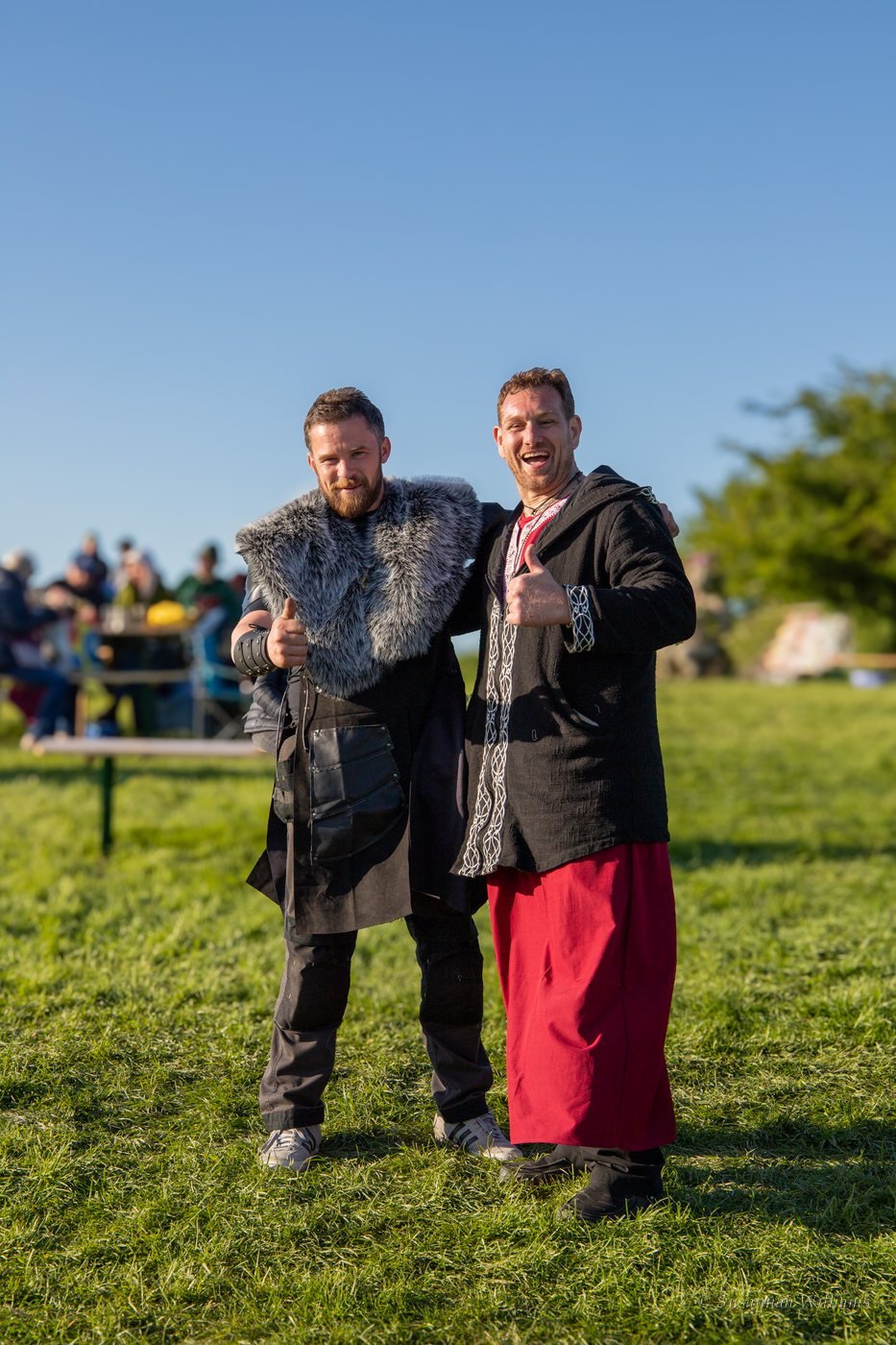 Two men in festival costume, one in a fur collar, both giving a thumbs up.