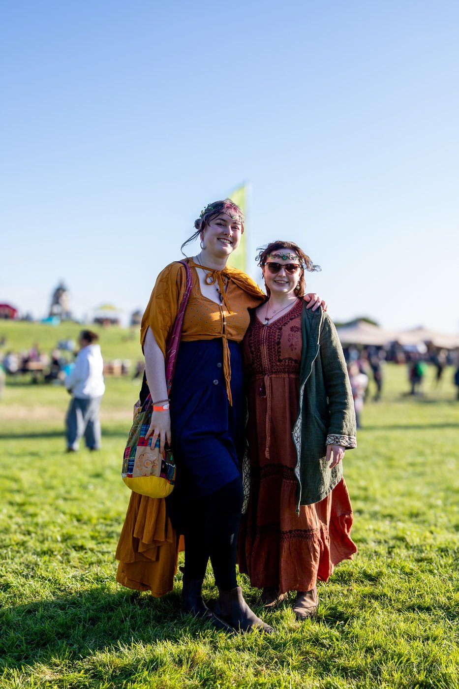 Two young women in flowing dresses and floral circlets, arm in arm on the hill.