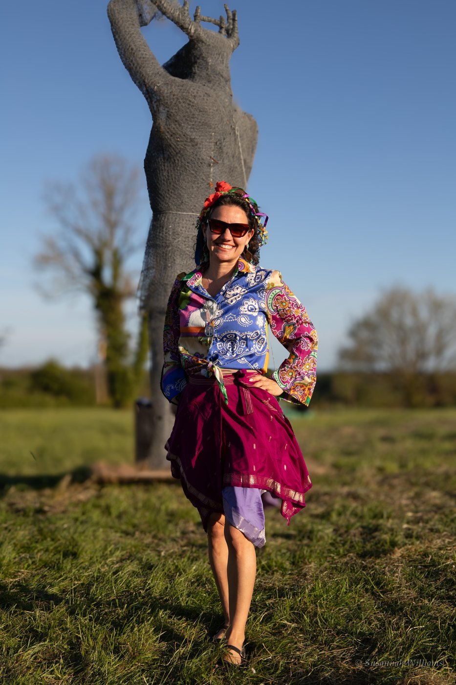A smiling woman in a patterned shirt and silk skirt in front of a large wire sculpture of a figure.
