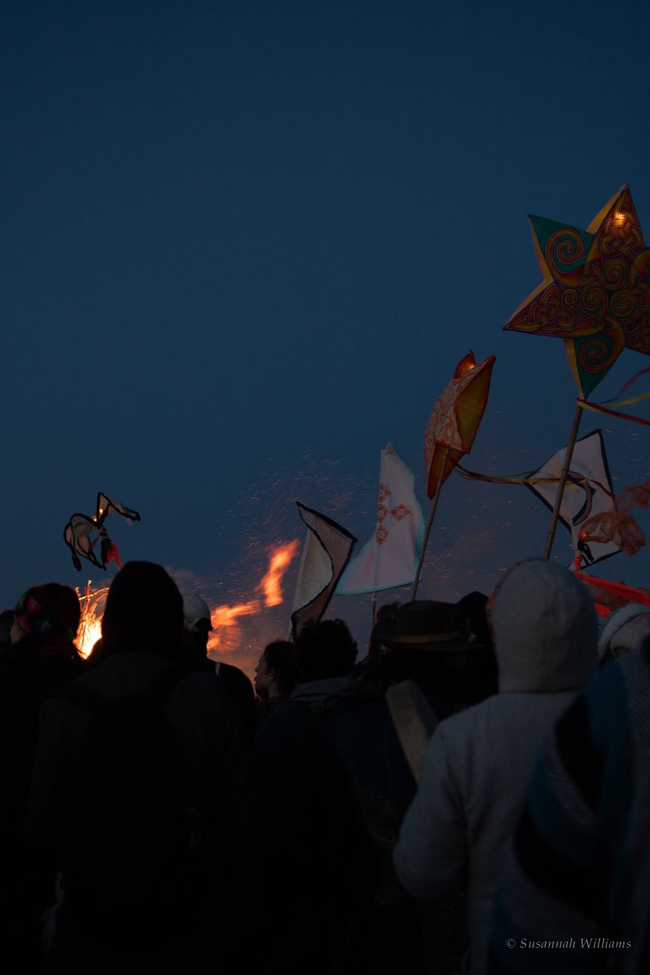 Silhouetted crowd beneath painted star lanterns and banners as sparks rise from the fire at night.