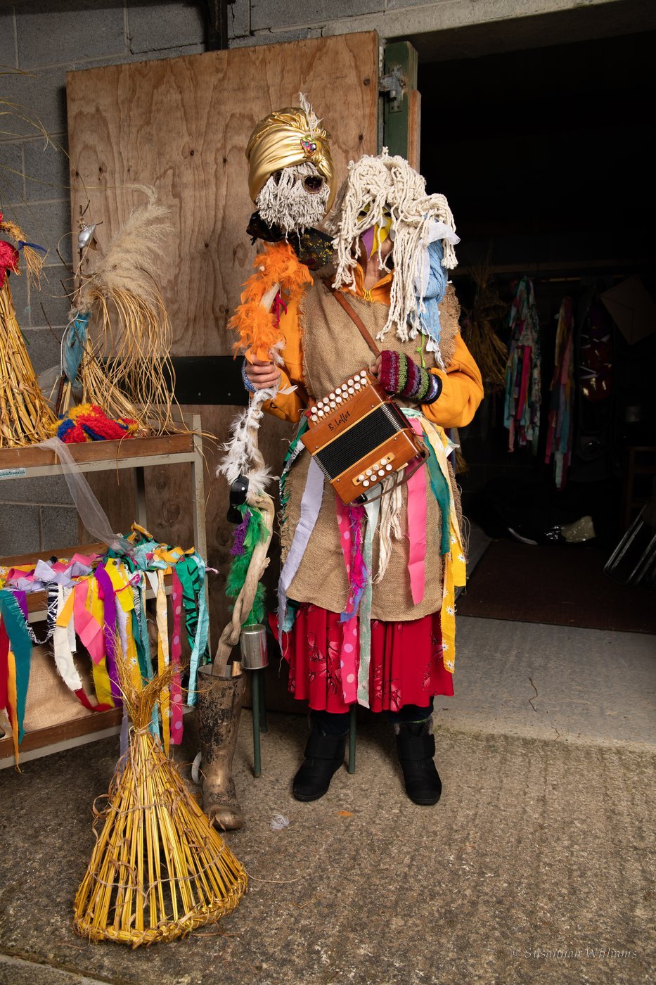 A Mummer in a mop-head mask and rag costume holding an accordion in a barn doorway, a straw hat on the ground beside her.