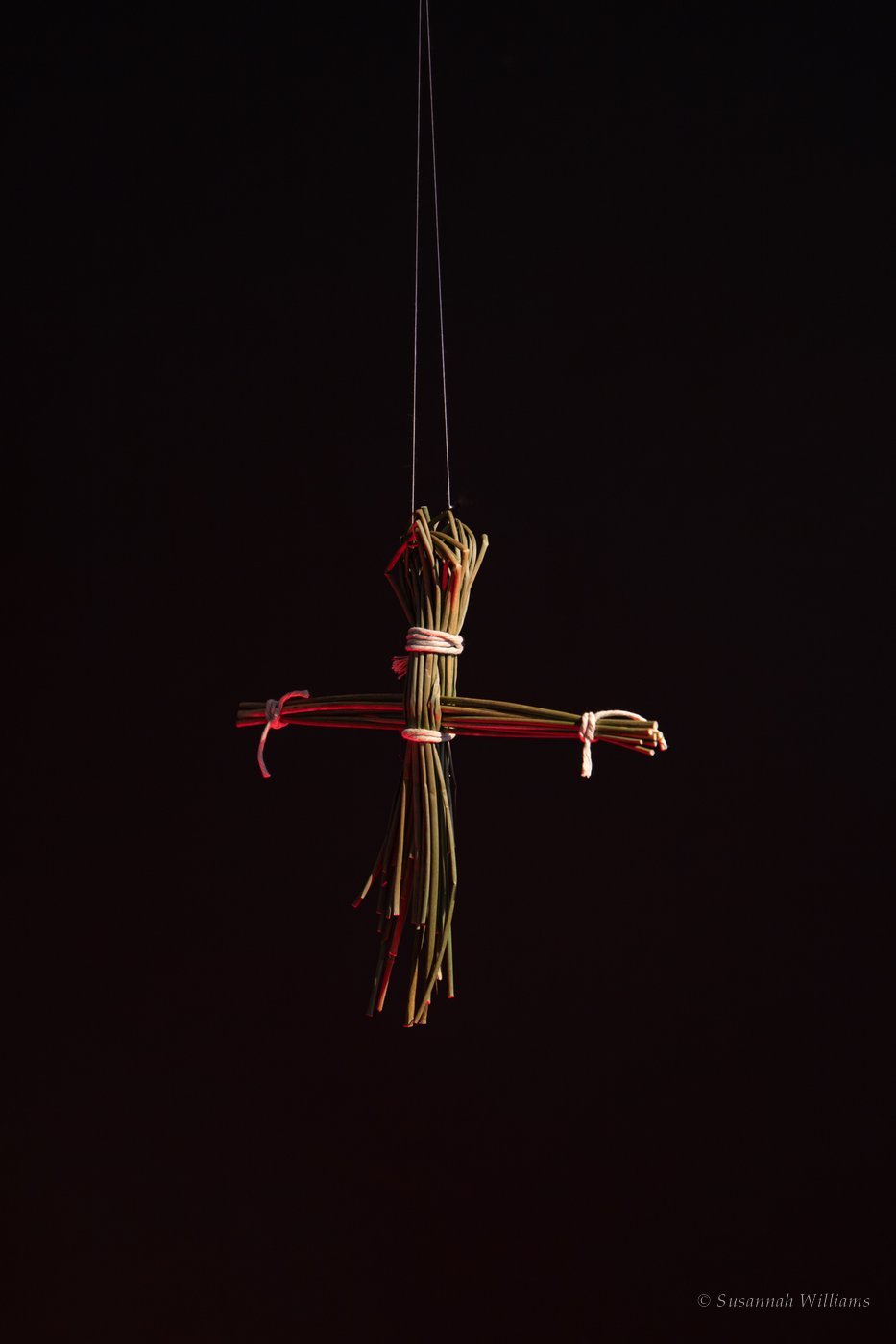 A Brigid’s cross made of rushes, hanging on fine thread against black.