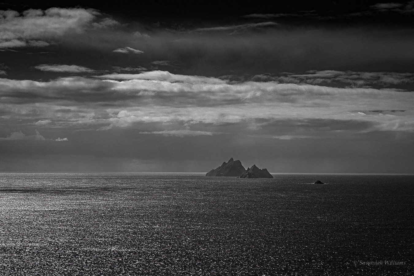 Black and white seascape: jagged island peaks on the horizon under heavy cloud, light glittering on the sea.