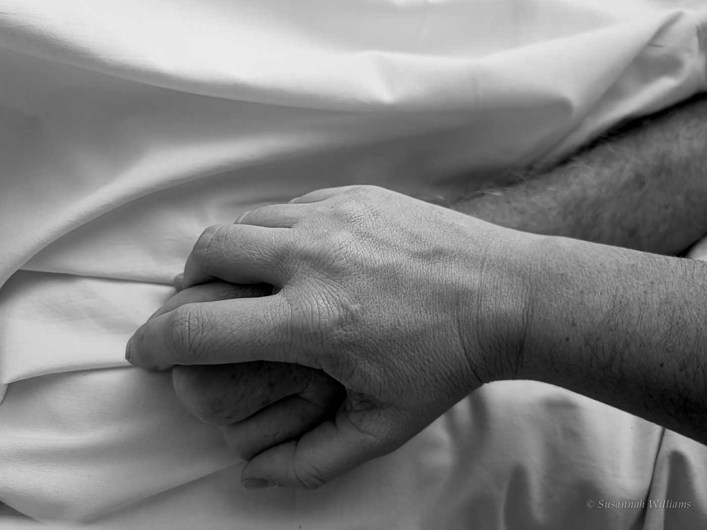 Black and white close-up of a hand holding another hand on a white hospital sheet.