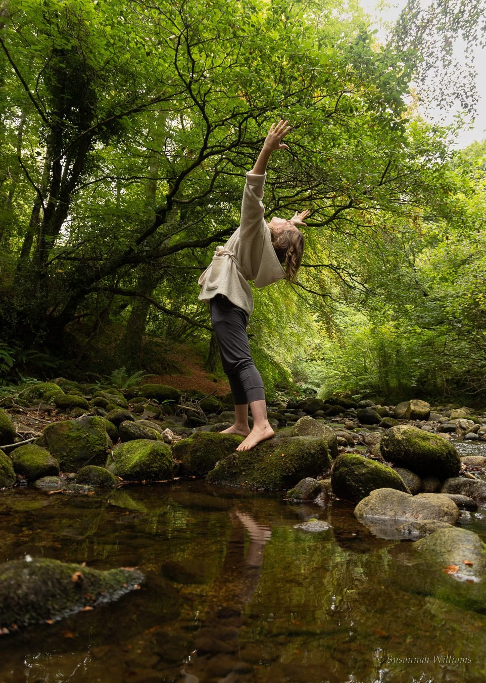 A woman barefoot on a mossy rock in a woodland river, reaching up towards the trees.