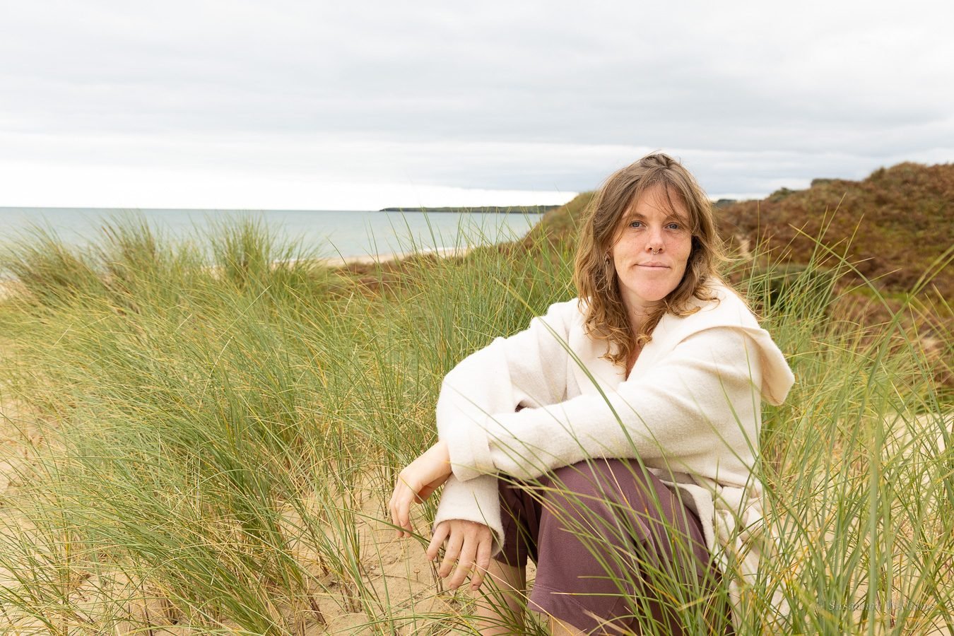 A woman in a cream wool coat sitting among marram grass in the dunes, the sea behind her.