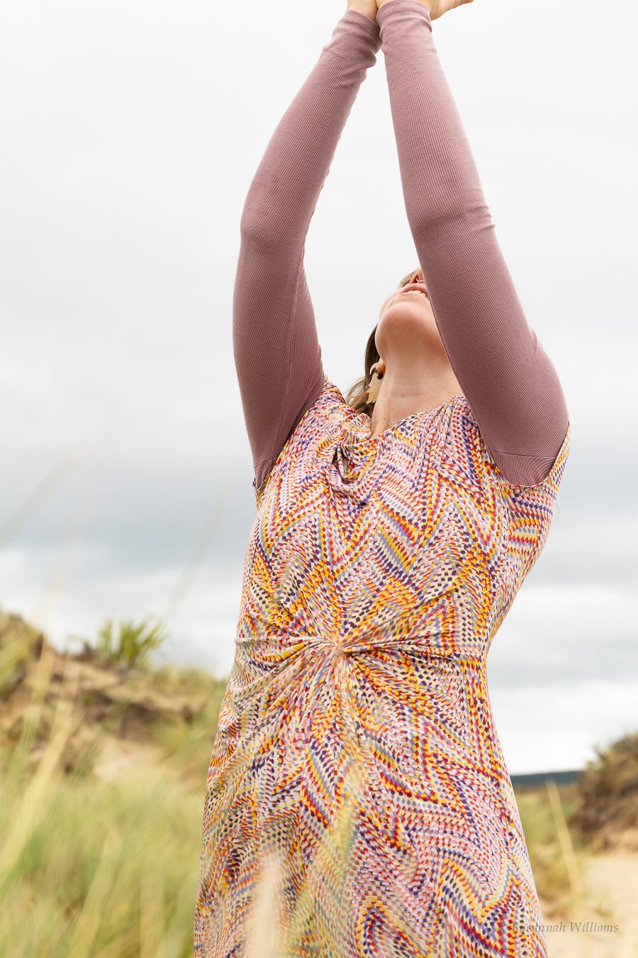 Arms raised overhead in a patterned dress against a grey sky.