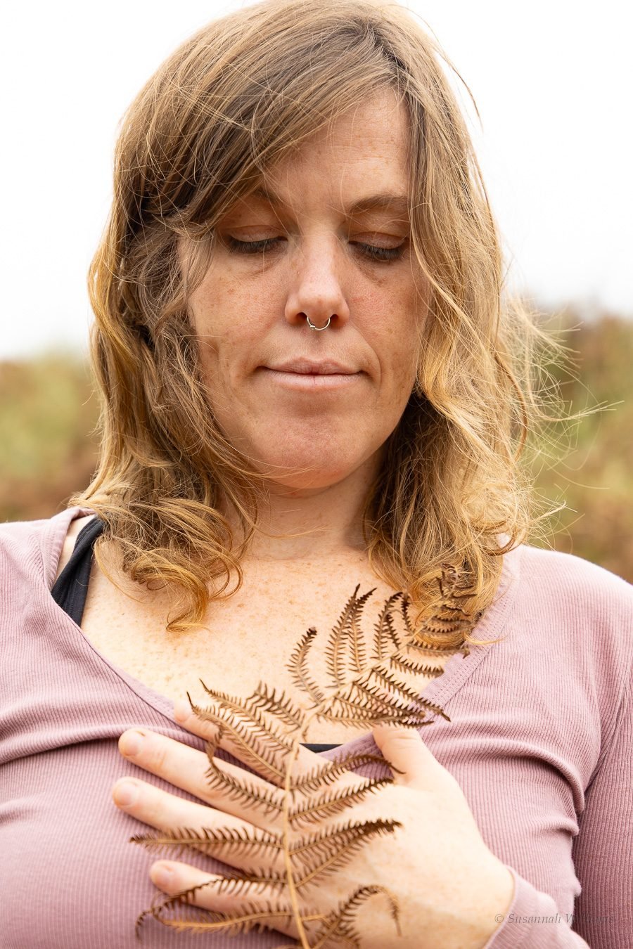 Eyes lowered, holding a dry fern frond against her chest.