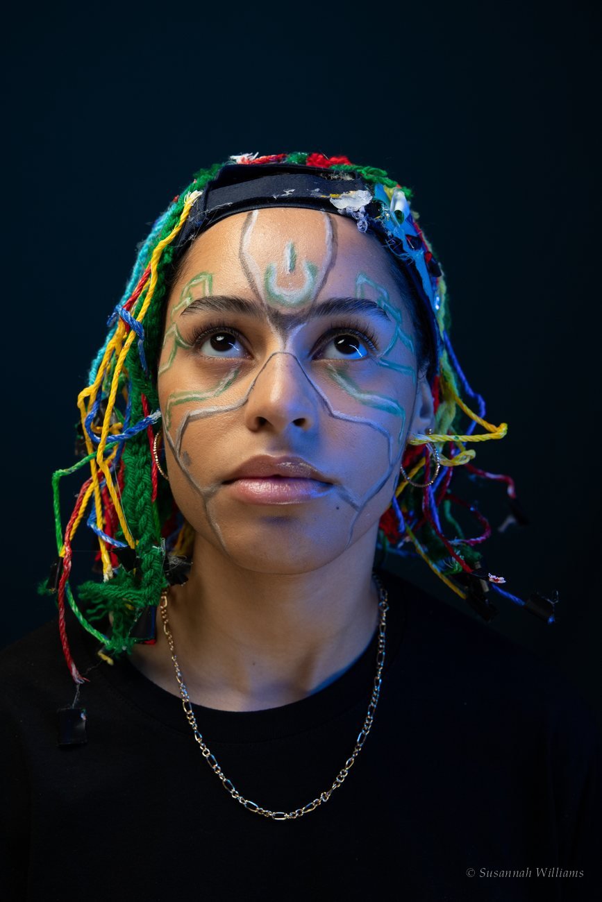 A woman with painted line markings on her face and braided coloured rope in her hair, looking upward.