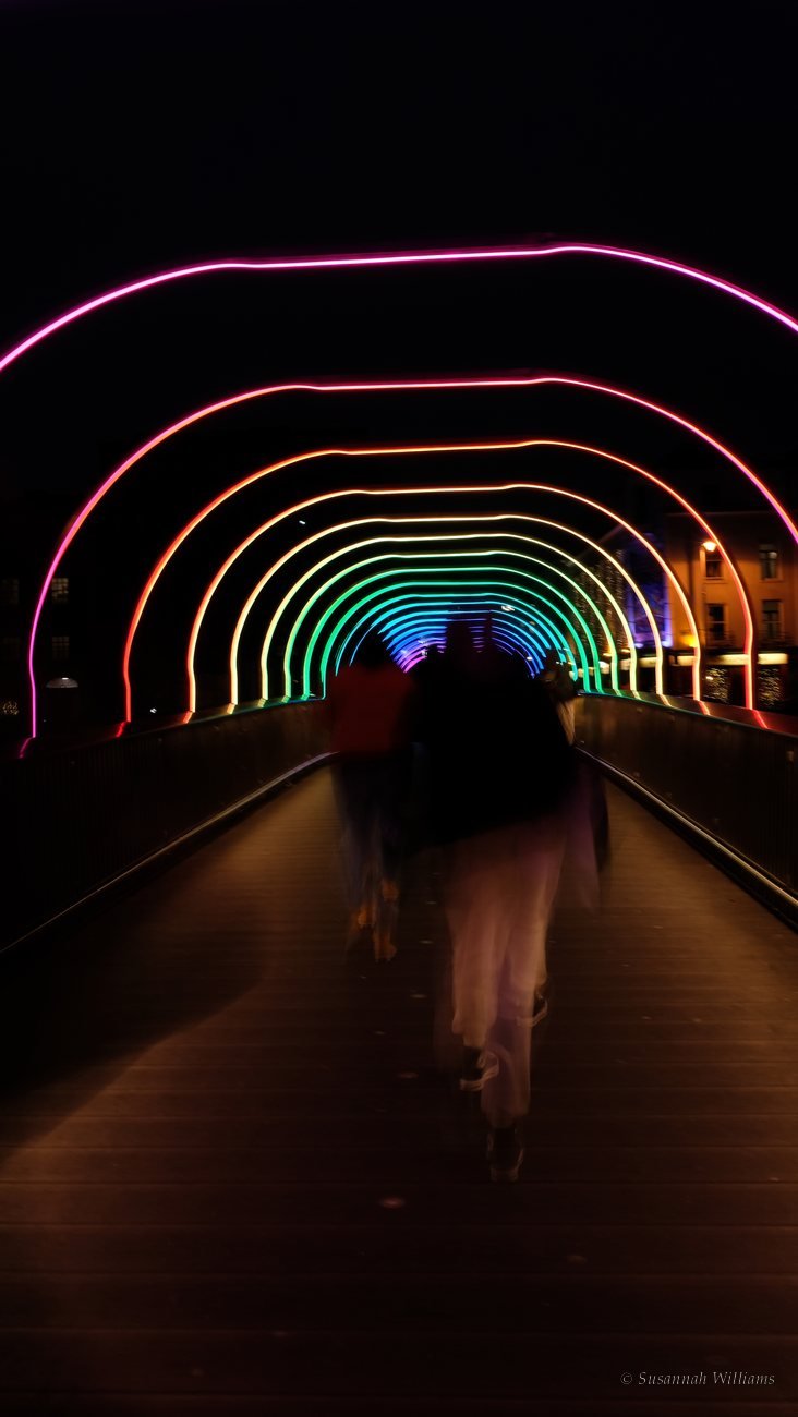 Blurred figures walking through rainbow-lit arches on a bridge at night.