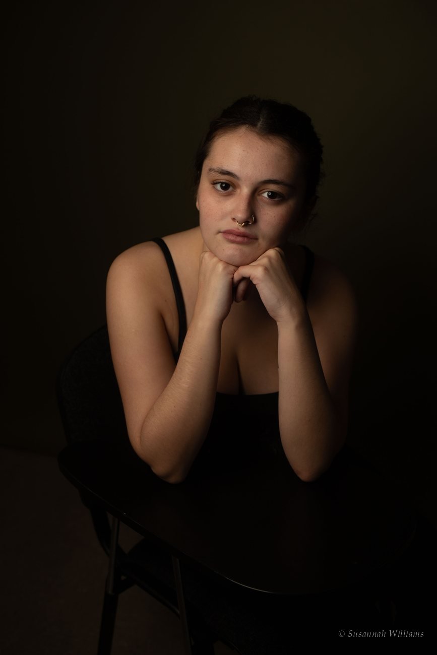 A young woman leaning on the back of a chair, chin resting on her fists, in warm low light.