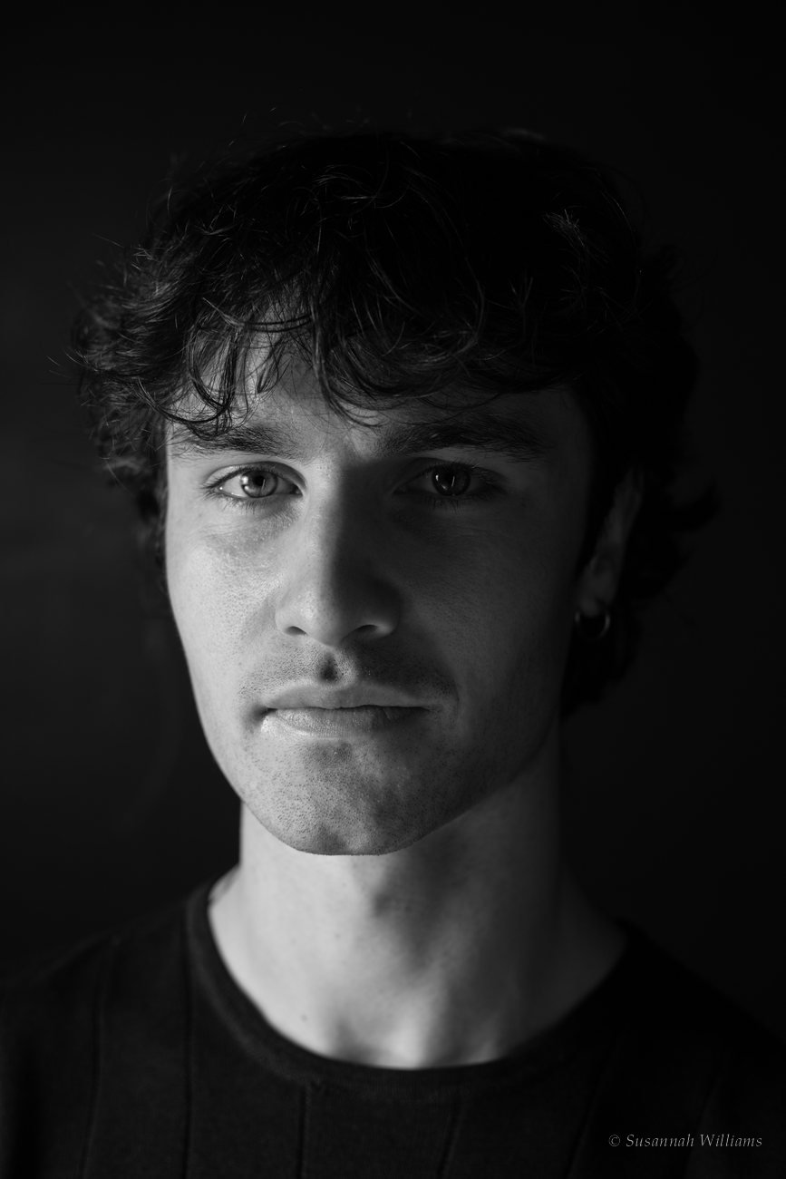 A black and white close portrait of a young man with dark curly hair, half his face in shadow.