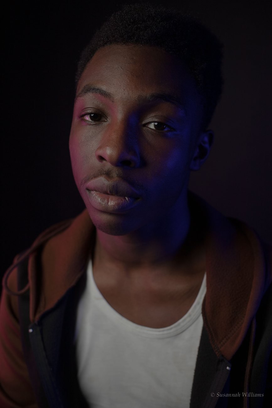 A young man in a white top and brown jacket, his face lit in pink and blue.