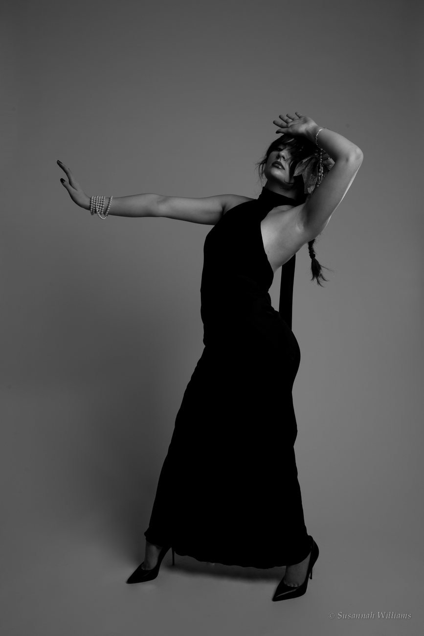 A black and white full-length portrait of a woman in a long black halter dress, arms extended in a dance pose.