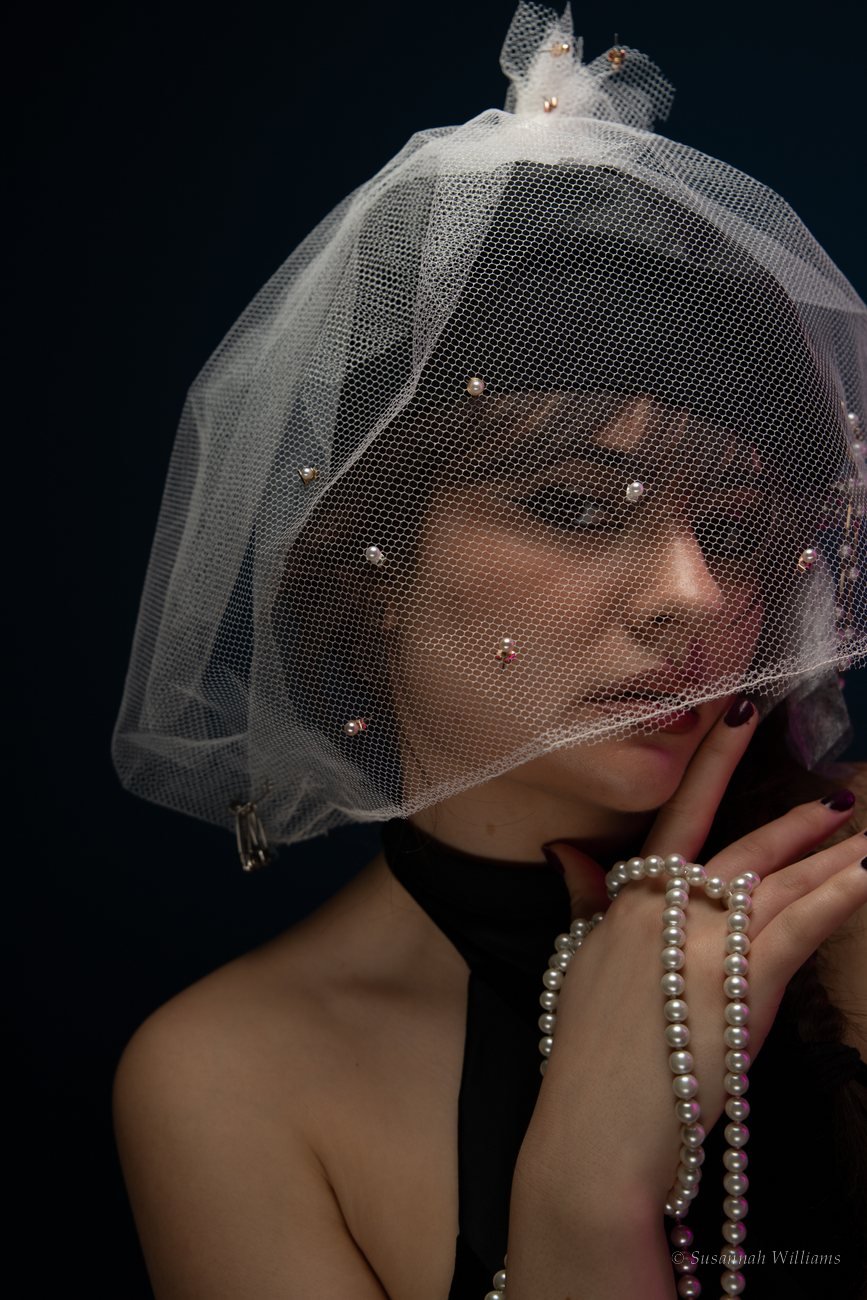 A woman in profile looking over her shoulder, wearing a black cap trimmed with pearls and torn fabric.
