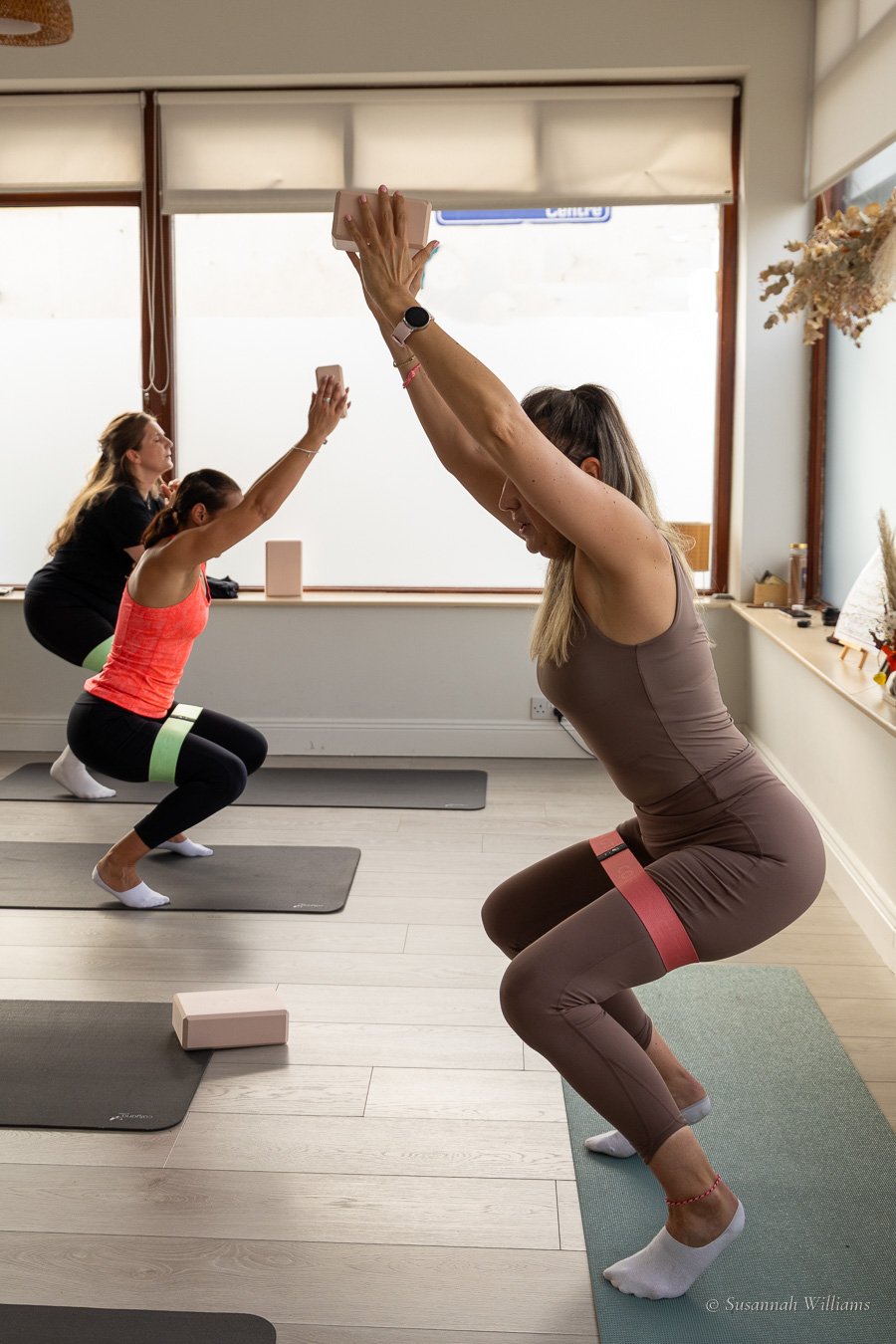 A Pilates teacher in a chair pose holding a block overhead, two women behind her copying the pose in front of a bright window.