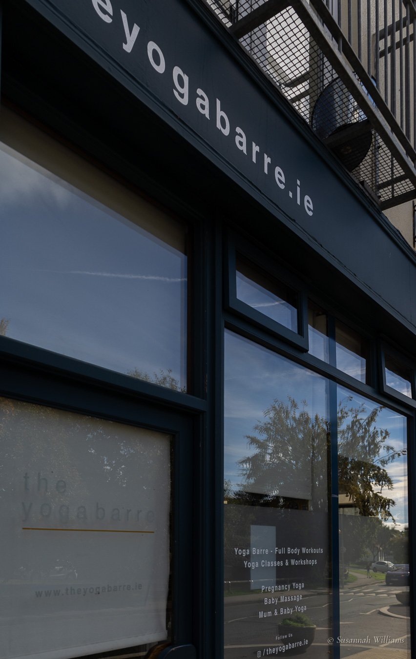 The navy shopfront of The Yoga Barre Studio, its name lettered on the frosted window and trees reflected in the glass.