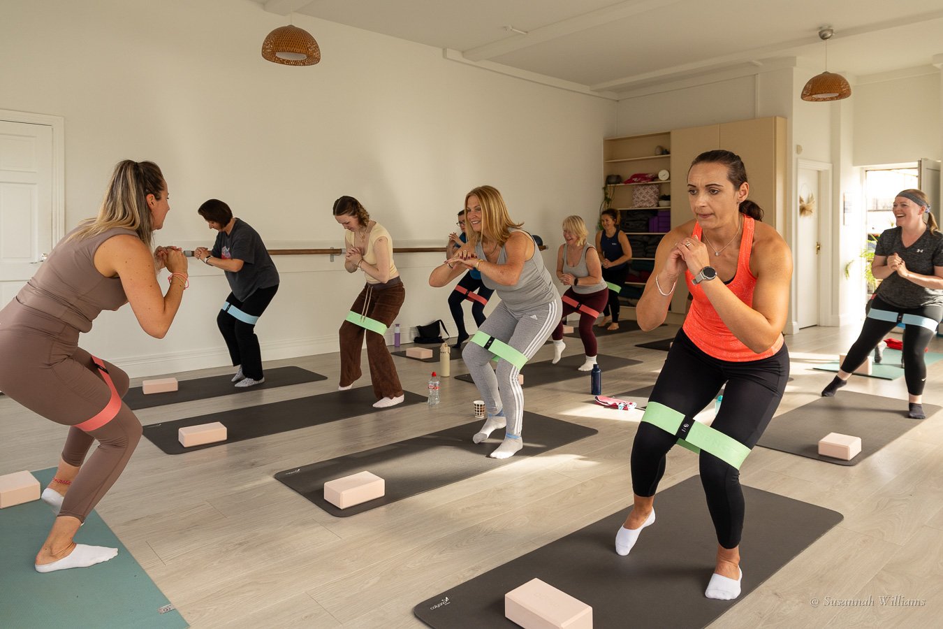 The teacher leading the class in a squat with resistance bands, a woman in orange in the foreground.