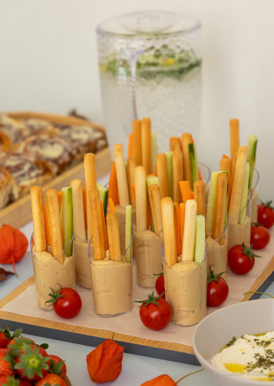 Cups of carrot and cucumber sticks with hummus, cherry tomatoes and a jug of infused water.
