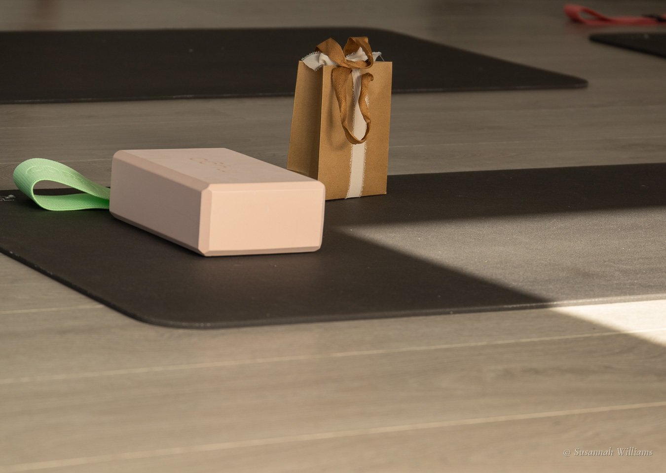A pink block, a green band and a small brown gift bag tied with ribbon on a mat in the sun.