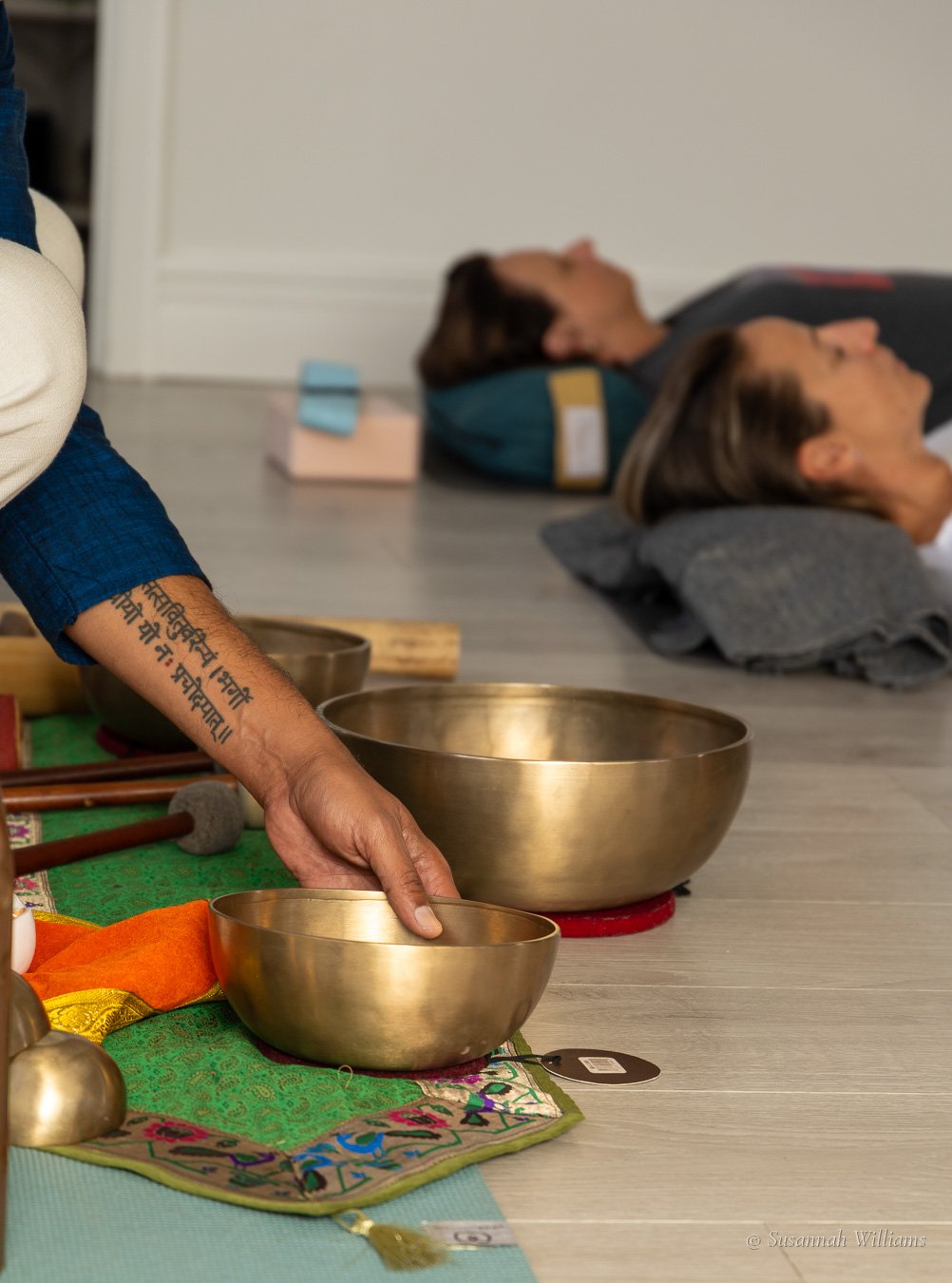 A tattooed hand on a singing bowl, people lying on mats behind.