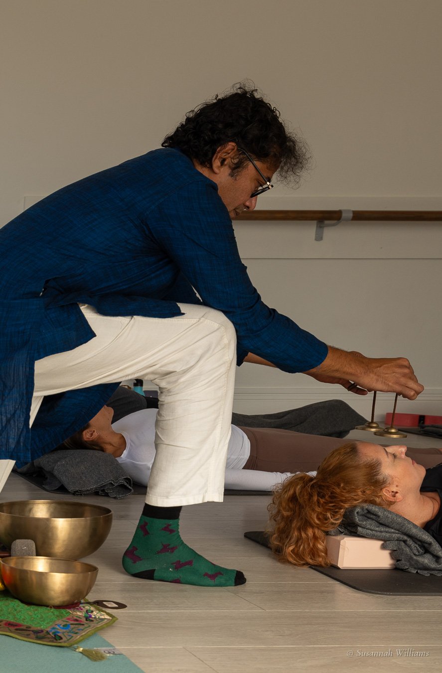 Parag leaning over a woman with red hair as she rests, ringing a pair of small brass chimes above her.