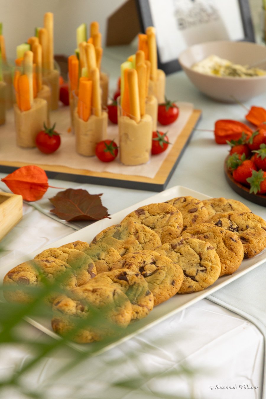 A tray of chocolate chip cookies seen through palm leaves, hummus cups and strawberries on the table behind.